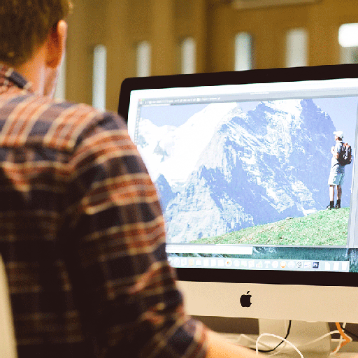 A person sitting at a desk with their back to the camera, looking at an Apple Mac screen, which shows a figure standing on grass with mountains in the background.
