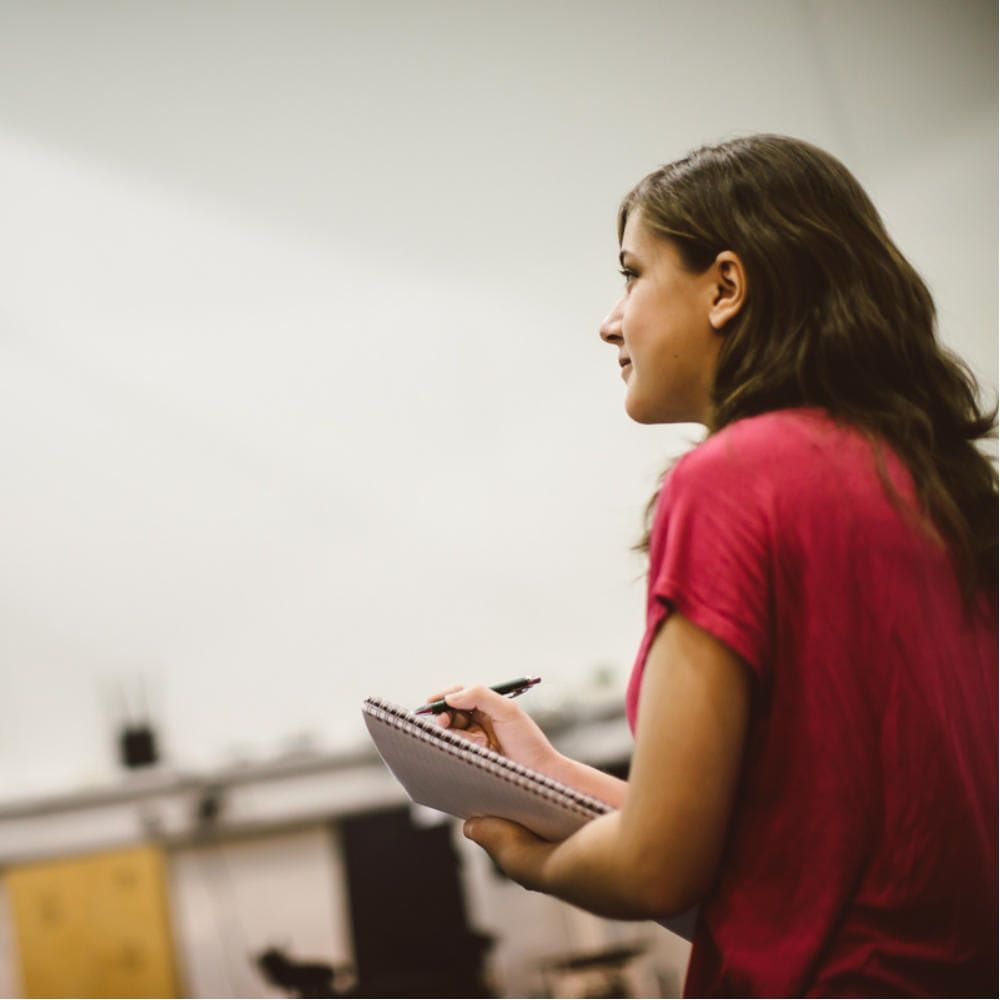 A student standing in a room looking up slightly, about to write in a notebook she's holding.