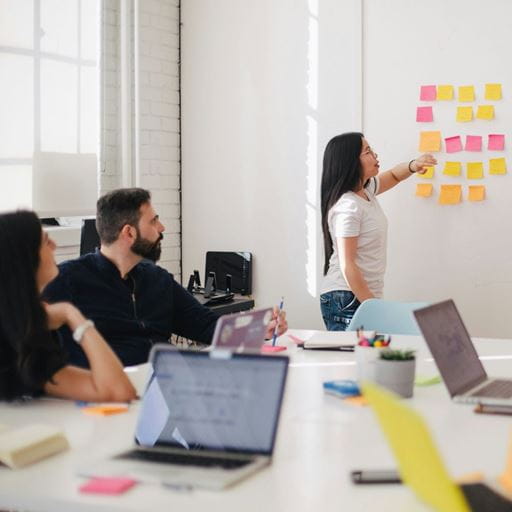 Image of a woman putting post-it notes on a whiteboard while two other people sit with laptops at a table watching her
