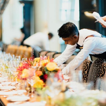 A man leaning over a table laid out for a meal with floral centrepieces and glassware in the foreground.