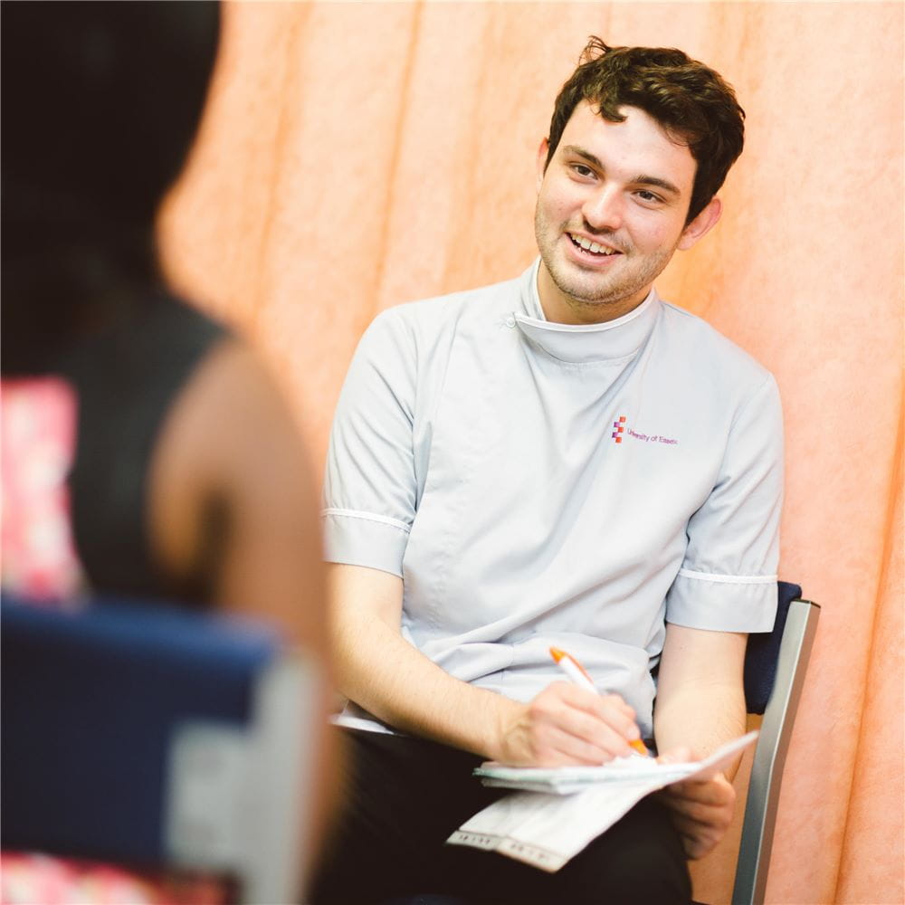 A nurse sitting with his back to some peach-coloured curtains, some paper on his lap and a pen in his hand. The photo is taken over the shoulder of a second person, who is in the foreground on the left of the photo.