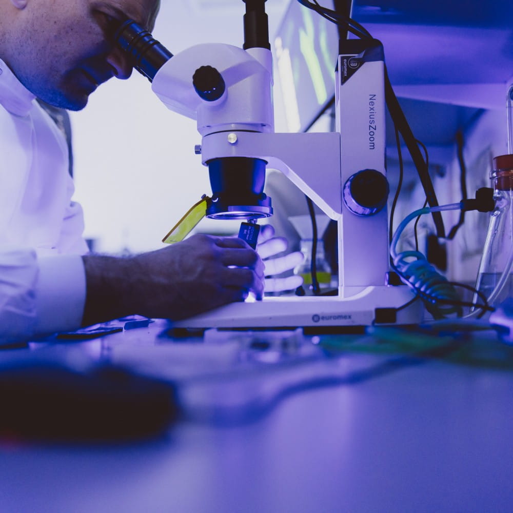 A blue-lit image of a person in a white lab coat looking down a microscope.