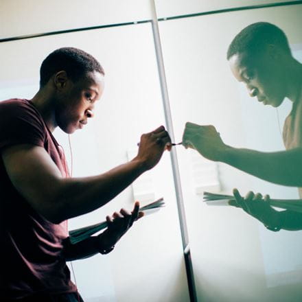 A student looking at a book, holding a pen up to write on a window. His reflection in the window is on the right.