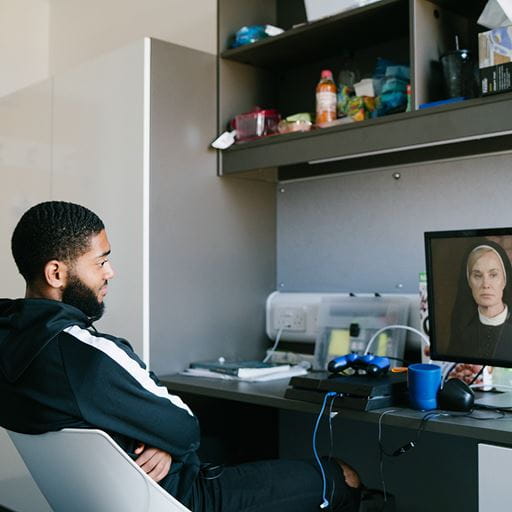 A young man sitting on a chair at a desk, watching something on a screen. Various items are also on the desk, and on some shelving above.