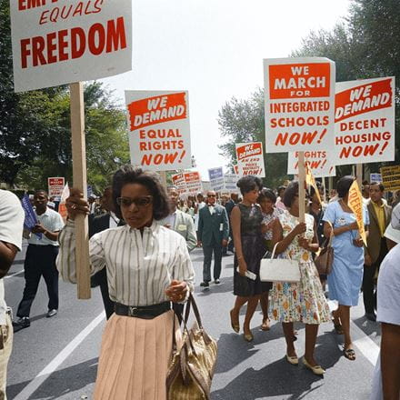 A small crowd of Black adults walking down a road carrying signs calling for the end of segregation in the USA.